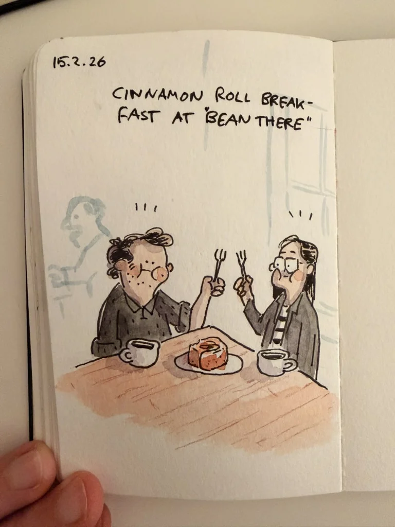 Marcel and Anna sit at a café table with cups and forks, ready to share a cinnamon roll for breakfast.