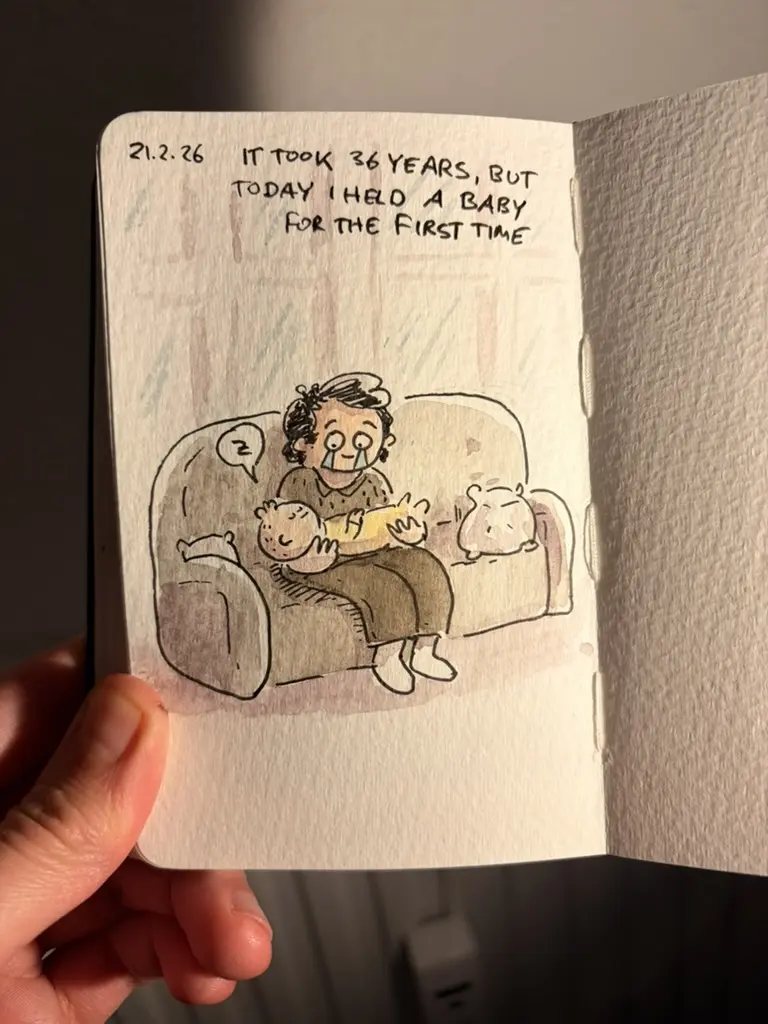 Marcel holds a baby for the first time while sitting on a sofa beside a cat.