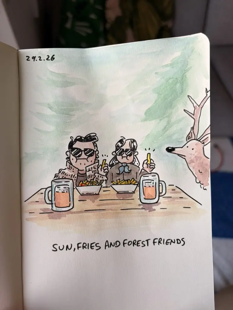Marcel and Anna eat fries and drink beer at a table in the forest as a deer looks on.